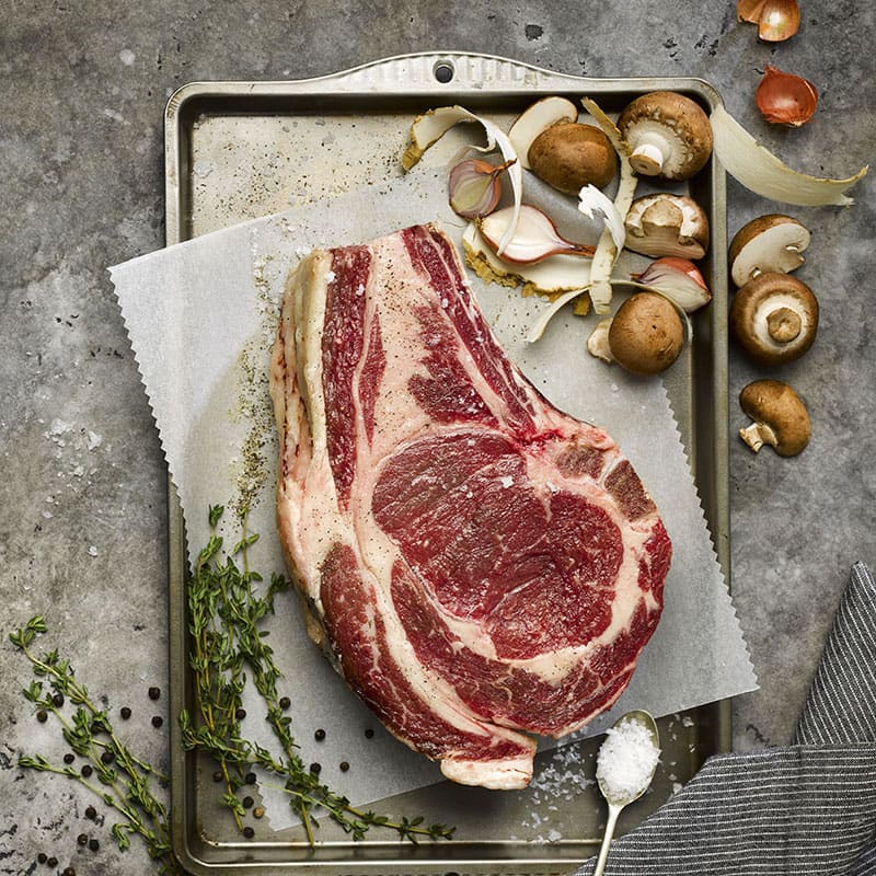 A raw Welsh dry aged beef rib joint on a wire rack, surrounded by fresh mushrooms and a sprig of rosemary, ready for cooking.